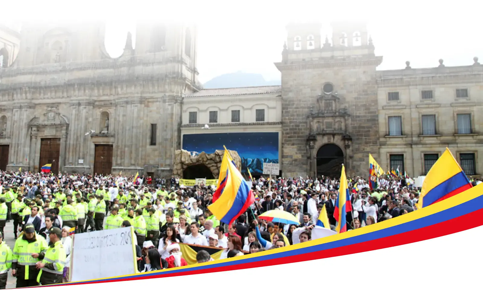 コロンビアに大勢の人々が集まり、その多くがコロンビアの国旗を掲げている。その多くはコロンビアの国旗を掲げている。おそらくは大聖堂か政府の建造物だろう。手前には法執行官がいる。